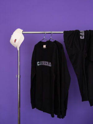 White baseball cap resting on a chrome clothing rack with black hoodies hanging against a purple background.