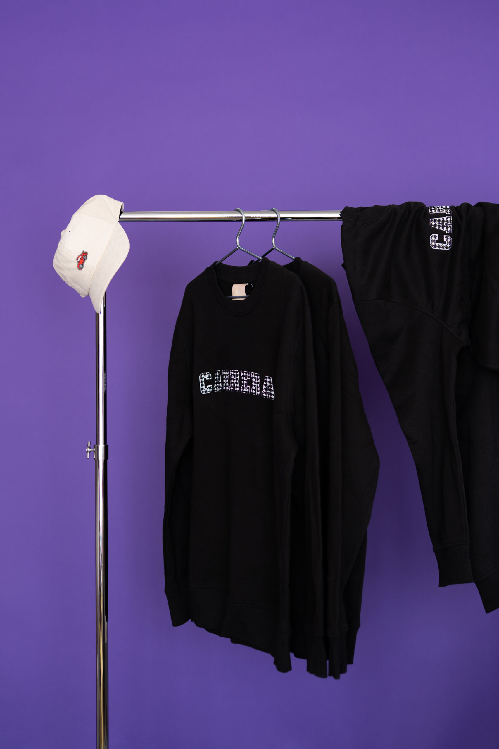 White baseball cap resting on a chrome clothing rack with black hoodies hanging against a purple background.