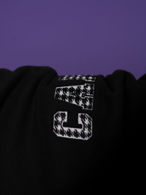 Close-up of a black hoodie with a white houndstooth-letter G patch on a purple background.