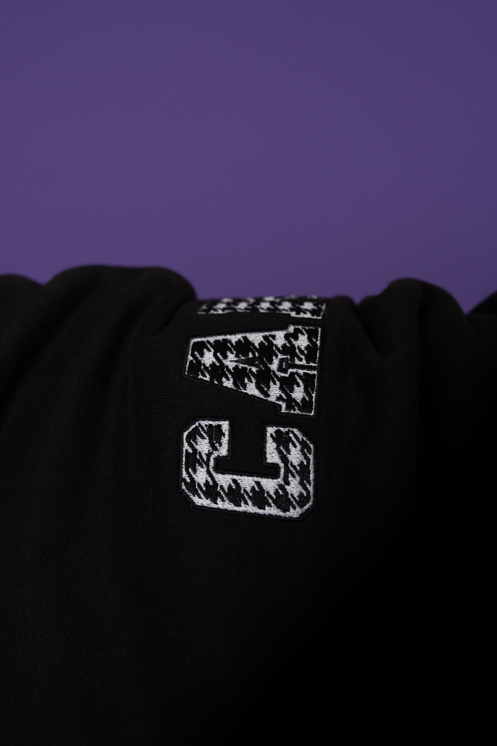 Close-up of a black hoodie with a white houndstooth-letter G patch on a purple background.