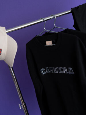 Black sweatshirt on a silver clothing rack against a purple background, with a white hat visible on the left.