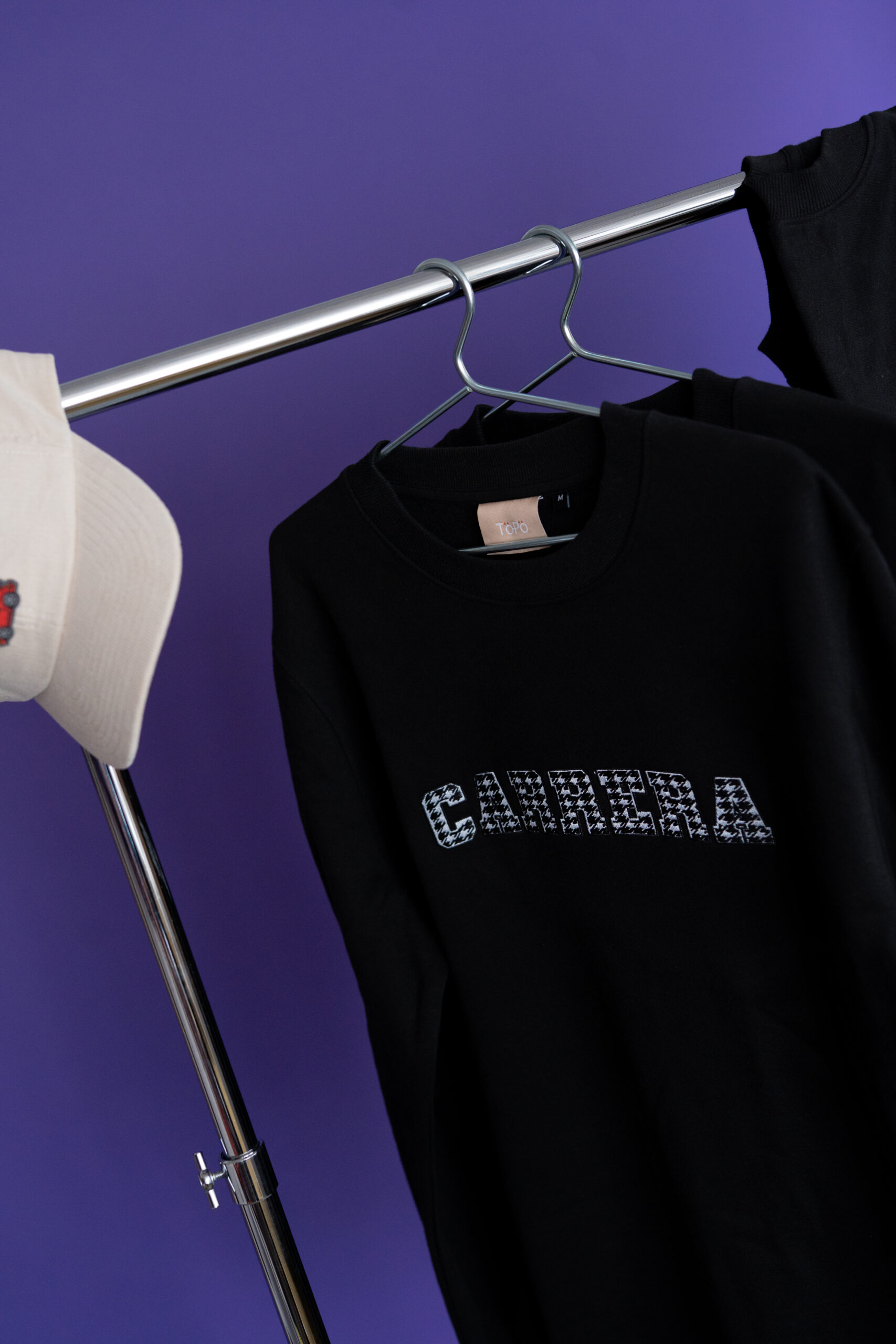 Black sweatshirt on a silver clothing rack against a purple background, with a white hat visible on the left.